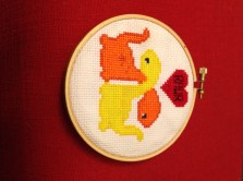 Cross-stitch of two dinosuars hugging with their necks. A red heart above their heads says "Rawr"