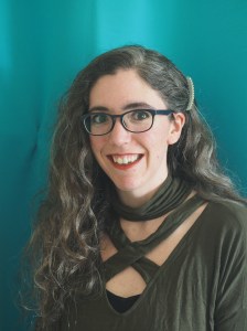 A white woman with curly brown hair stands in front of a blue backdrop. She's wearing glasses and a green top and is smiling.