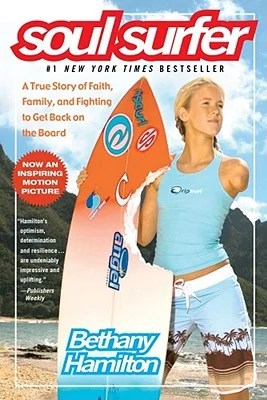A white woman with one arm holds a surf board with a bite taken out of it.